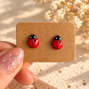 May include: A pair of stud earrings shaped like ladybugs with bright red wings, black spots, and black heads. The earrings are attached to a rectangular brown cardboard display card, showcasing the detailed insect design.