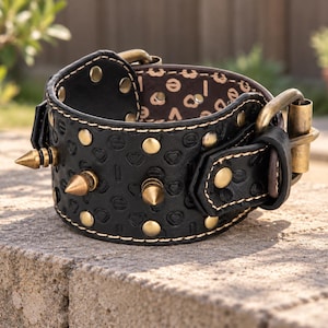 May include: A wide black leather cuff bracelet featuring a repeating embossed heart pattern, brass-toned conical spikes, and round metal studs. The bracelet includes a brass-toned buckle closure and a dark brown interior lining with a repeating heart and letter print.