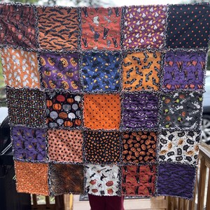 May include: A square rag quilt featuring a patchwork of twenty-five fabric squares with various Halloween-themed prints including bats, pumpkins, ghosts, skeletons, and cats in orange, purple, black, and white colors with frayed seams.