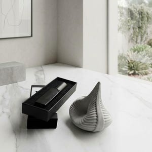 May include: A matte black rectangular perfume bottle rests inside a black box, accompanied by a light gray, spiraled, teardrop-shaped ceramic sculpture. The items are arranged together, showcasing a contrast between the dark, angular packaging and the light, textured, organic form of the sculpture.