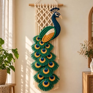 May include: A macrame wall hanging featuring a peacock design in shades of deep blue, emerald green, and golden yellow. The peacock is centered on a cream-colored knotted cord background suspended from a horizontal wooden dowel rod.