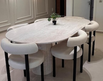 Puede incluir: Una mesa de comedor ovalada con tablero de mármol beige claro y base cilíndrica, rodeada de seis sillas con asientos tapizados en color crema y respaldos curvos sobre patas negras. El texto MARBRE DE CLASSE es visible en la superficie de la mesa.