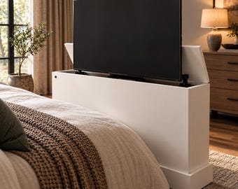 May include: A white rectangular television lift cabinet featuring a motorized mechanism that extends a flat screen television upward from the interior. The unit has a smooth finish and is positioned at the foot of a bed frame.