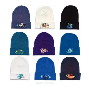 May include: A collection of nine knit beanies in various colors including navy, white, black, teal, purple, and blue. Each beanie features a unique embroidered graphic of a stylized animal or creature centered on the folded cuff.