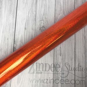 May include: A roll of orange glitter vinyl with a metallic sheen. The vinyl is rolled up and the end is visible. The vinyl is on a wood background.