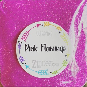 May include: A close-up of a bright pink, glittery surface with a circular label. The label reads "Pink Flamingo" in black text, with the word "ULTRAFINE" above it. The website "Zindee.com" is also visible.