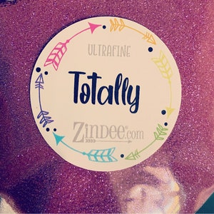May include: A round label with a white background and a pink glitter texture. The label features a circle of colorful arrows with the text "ULTRAFINE Totally Zindee.com" in the center.