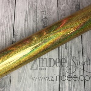 May include: A roll of gold glitter vinyl with a holographic finish. The vinyl is rolled up and has a slight curve to it. The vinyl is on a wooden surface.