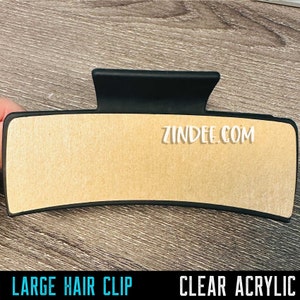 May include: A large black plastic hair clip with a clear acrylic top. The clip has a textured surface and the text "zindee.com" is printed on the top.