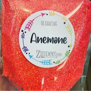 May include: A close-up of a bag of ultra-fine orange glitter labeled "Anemone" with a white circle sticker with black arrows and the text "Zindee.com".