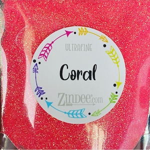 May include: A bag of ultrafine coral glitter. The glitter is a vibrant, sparkling coral color. A white circular label with the word "Coral" in black script is centered on the bag. The label also includes the words "ULTRAFINE" and "Zindee.com".