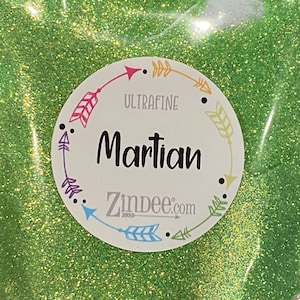 May include: Close-up of a bag of green glitter labeled "Martian" with the text "Ultrafine" and "Zindee.com" on a circular sticker. The glitter is a bright, sparkling green.