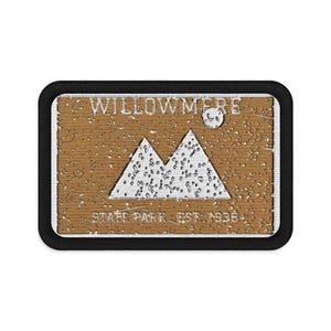 Puede incluir: Un parche rectangular bordado con un borde negro. El centro presenta un fondo marrón con un gráfico de montaña blanco, un círculo blanco que representa el sol y el texto WILLOWMERE STATE PARK EST. 1938 en letras blancas.