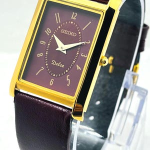 May include: A rectangular Seiko Dolce wristwatch featuring a gold-tone case and a deep burgundy dial. The face displays gold-colored hour markers, hands, and the text Seiko and Dolce. It is paired with a matching dark burgundy leather strap.