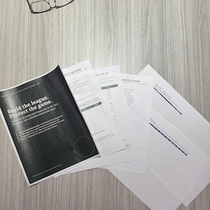 May include: A stack of printed documents featuring a black cover page with the white text Build the league. Protect the game. and additional pages with tables and lists, accompanied by a pair of black-rimmed eyeglasses resting nearby.