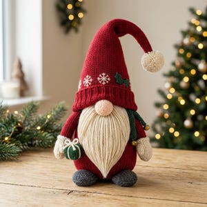 Könnte beinhalten: Eine Plüsch-Wichtelfigur mit einer hohen, roten Strickmütze, die mit weißen Schneeflocken bestickt ist und eine Bommelspitze hat. Der Wichtel besitzt einen langen, cremefarbenen Garnbart, eine runde Nase und Strickfäustlinge.