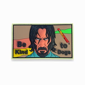 May include: A rectangular PVC morale patch featuring a stylized illustration of a man with long dark hair and a beard. The text "Be Kind to Dogs" is printed on the patch, which also includes a graphic of a yellow pencil with a red eraser.