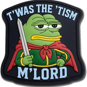 May include: A black rubber patch featuring a green cartoon frog character wearing a red cape and holding a silver sword. The text T'WAS THE 'TISM M'LORD is printed in light blue letters at the top and bottom of the shield-shaped design.