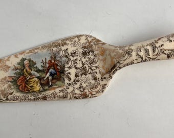 Könnte beinhalten: Ein Tortenheber aus Keramik mit cremefarbener Basis, verziert mit einem goldenen Blumenmuster und einer zentralen Abbildung von zwei Figuren in einer Landschaft. Der Griff hat ein Loch zum Aufhängen und eine strukturierte Grifffläche.