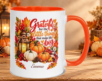 May include: A white ceramic mug with an orange interior and handle, featuring a colorful autumn-themed graphic of pumpkins, a lantern, and fall foliage. The text reads Grateful for the little things that make life big and Emma.