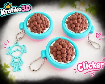 Puede incluir: Tres clickers con forma de taza en turquesa y rosa con esferas texturizadas marrones que parecen cereales, cada uno unido a una cadena de bolas plateada. Un clicker incluye un dije turquesa con forma de persona. Texto Krafiko3D y Clicker.