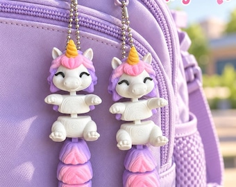 Puede incluir: Dos llaveros de unicornio articulados con cuerpos blancos, crines rosas y moradas y cuernos amarillos. Los llaveros cuentan con partes segmentadas móviles y están sujetos a una mochila morada clara con cadenas de bolas plateadas. Texto: Krafiko3D Unicorn Keychains.