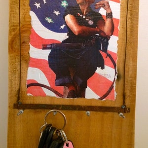 May include: A wooden sign with a distressed American flag background featuring a woman in a blue jumpsuit holding a wrench. The sign has a metal key holder with a set of keys attached.