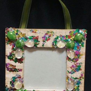 May include: A small wooden picture frame decorated with colorful confetti and green and white glass gems. The frame has a ribbon attached for hanging.