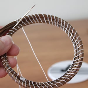 Może przedstawiać: Zbliżenie na trwający projekt rzemieślniczy, przedstawiający okrągły, tkany wzór. Brązowy i biały materiał jest zszywany igłą i białą nicią. Projekt jest trzymany przez rękę.