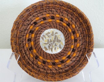 May include: A circular woven basket with a hollow center filled with reflective, irregular glass shards. The basket features concentric rings of brown pine needles, accented by vibrant orange and dark brown ribbon stitching throughout.