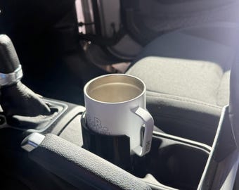 May include: A white stainless steel travel mug with a handle sits in a black plastic cup holder located in a vehicle centre console. The mug features a partial graphic print and the text MiiR printed vertically on the side of the handle.