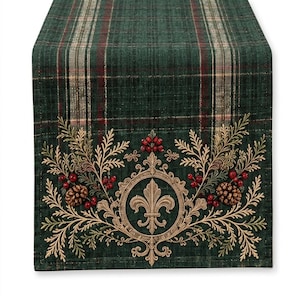 May include: A rectangular table runner featuring a dark green plaid pattern with thin red and beige stripes. The end is decorated with an embroidered gold fleur-de-lis emblem, pine branches, pinecones, and clusters of small red berries.