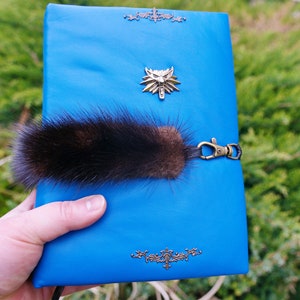 May include: A blue leather journal with a wolf emblem and a brown fur tail keychain. The journal has decorative gold accents.