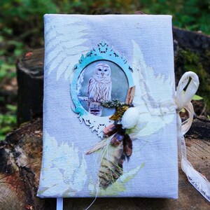 May include: A light gray journal with an owl illustration in a decorative frame. The cover features fern and floral accents, with a cream-colored ribbon tie. The journal is embellished with natural elements.