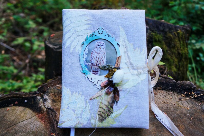 May include: A light gray journal with an owl illustration in a decorative frame. The cover features fern and floral accents, with a cream-colored ribbon tie. The journal is embellished with natural elements.