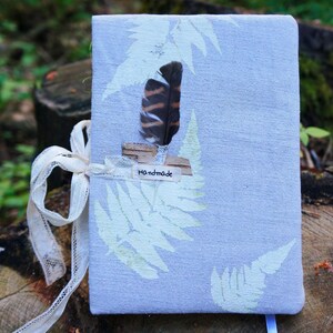 May include: A handmade journal with a light blue cover featuring fern leaf designs. A decorative feather and the word "handmade" are attached. The journal is tied with a cream-colored ribbon and sits on a tree stump.