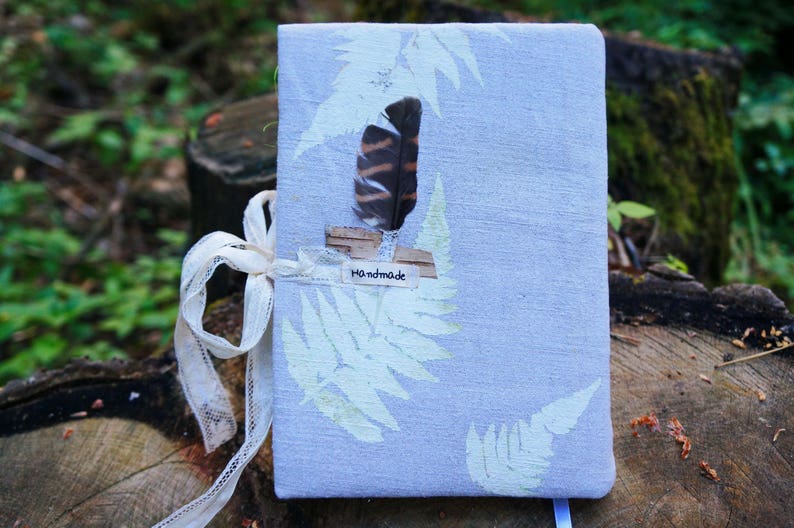 May include: A handmade journal with a light blue cover featuring fern leaf designs. A decorative feather and the word "handmade" are attached. The journal is tied with a cream-colored ribbon and sits on a tree stump.