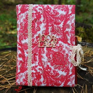 Pu&ograve; includere: Un taccuino con motivo paisley rosso e bianco con un bordo in pizzo color crema e un'etichetta in legno che dice "100% Lady".