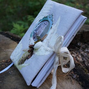 May include: A handmade journal with a light blue linen cover, adorned with an owl illustration in a turquoise frame. The journal is embellished with a white ribbon and natural elements like pinecones and leaves, suggesting a nature-inspired theme.