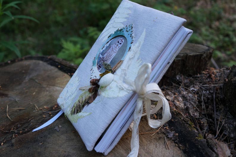 May include: A handmade journal with a light blue linen cover, adorned with an owl illustration in a turquoise frame. The journal is embellished with a white ribbon and natural elements like pinecones and leaves, suggesting a nature-inspired theme.
