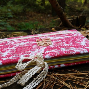Pu&ograve; includere: Un diario con motivi rosa e bianchi con un bordo in pizzo e un'etichetta in legno che dice "Do It". Il diario &egrave; chiuso e si trova su un letto di aghi di pino.