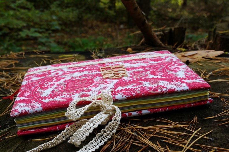 Pu&ograve; includere: Un diario con motivi rosa e bianchi con un bordo in pizzo e un'etichetta in legno che dice "Do It". Il diario &egrave; chiuso e si trova su un letto di aghi di pino.