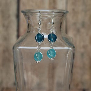 May include: A pair of drop earrings featuring two stacked beads each, wrapped in silver-toned wire. The top beads are a deep navy blue, while the bottom beads are a lighter turquoise blue. The earrings hang from silver-toned metal hooks.