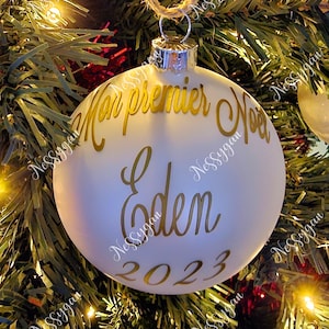 Peut inclure: Une boule de Noël blanche avec l'inscription dorée "Mon premier Noël Eden 2023". L'ornement est suspendu par une ficelle en corde naturelle, sur fond de sapin de Noël.