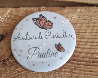 May include: A white round button with a gray background and orange butterflies. The text on the button reads "Auxiliaire de Puériculture Pauline".