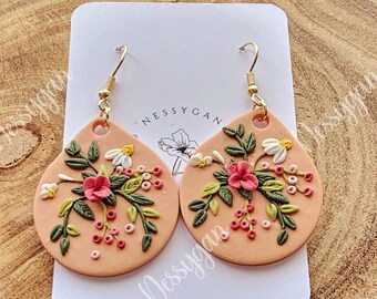 May include: A pair of peach-colored clay earrings with a floral design. The earrings feature pink and white flowers, green leaves, and small red berries. The earrings are on a white card with the word "NESSYGAN" written on it.