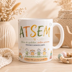 Peut inclure: Mug blanc avec le mot "ATSEM" en lettrage aquarelle orange et vert. La tasse présente un dessin d'enfants se tenant la main et du texte en français. Un cœur et un arc-en-ciel sont également inclus dans le design.