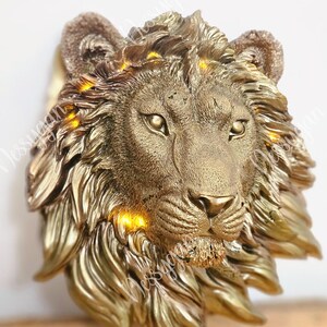 Op de afbeelding: Een gouden leeuwenkop wanddecoratie met gedetailleerde manen en gezichtskenmerken. De sculptuur heeft een getextureerde afwerking en is verlicht met kleine, warmgekleurde lampjes. De blik van de leeuw is naar voren gericht, en het ontwerp is koninklijk.