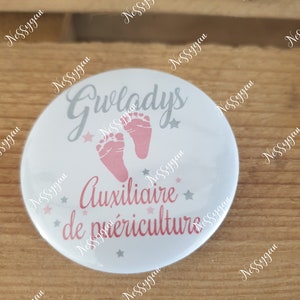 Peut inclure: Bouton blanc avec un design rose et gris. Le bouton dit "Gwladys Auxiliaire de puériculture" avec une paire d'empreintes de pieds de bébé roses et des étoiles.