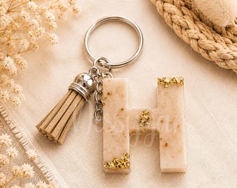May include: A letter H keychain made of beige resin with embedded gold flakes, featuring a silver-toned metal keyring and a matching beige faux suede tassel. The letter has a smooth, semi-translucent finish with gold leaf accents at the corners.